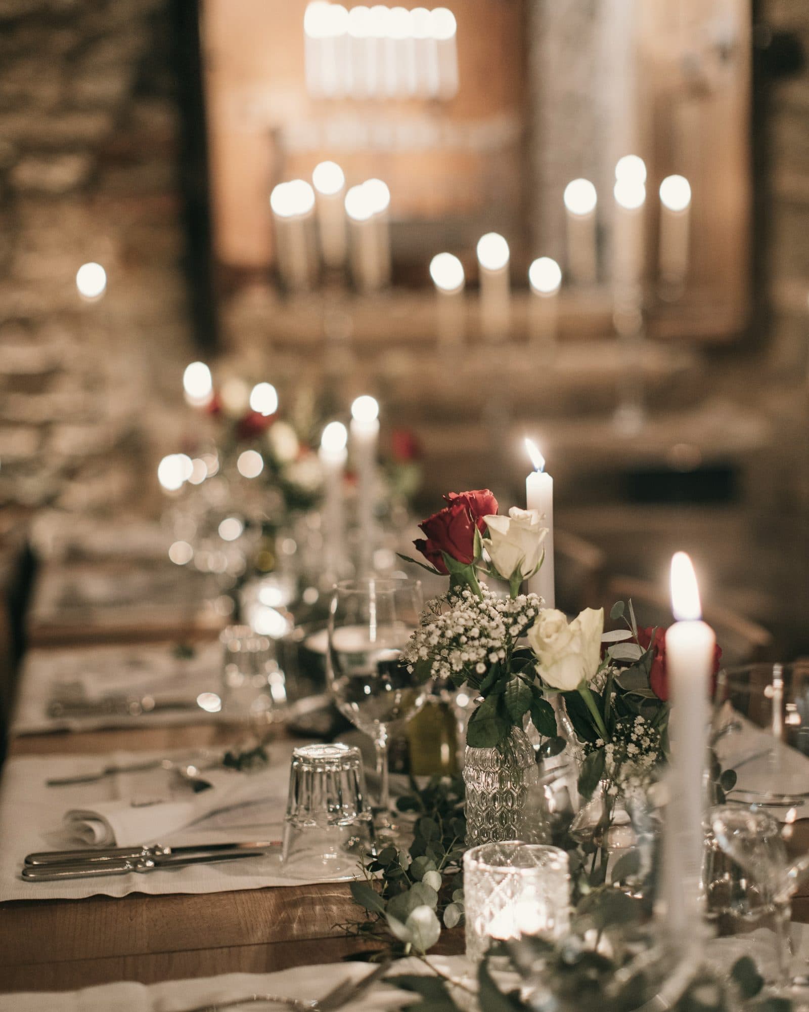 Mesa de banquete decorada con velas encendidas, rosas rojas y blancas, y copas de cristal.