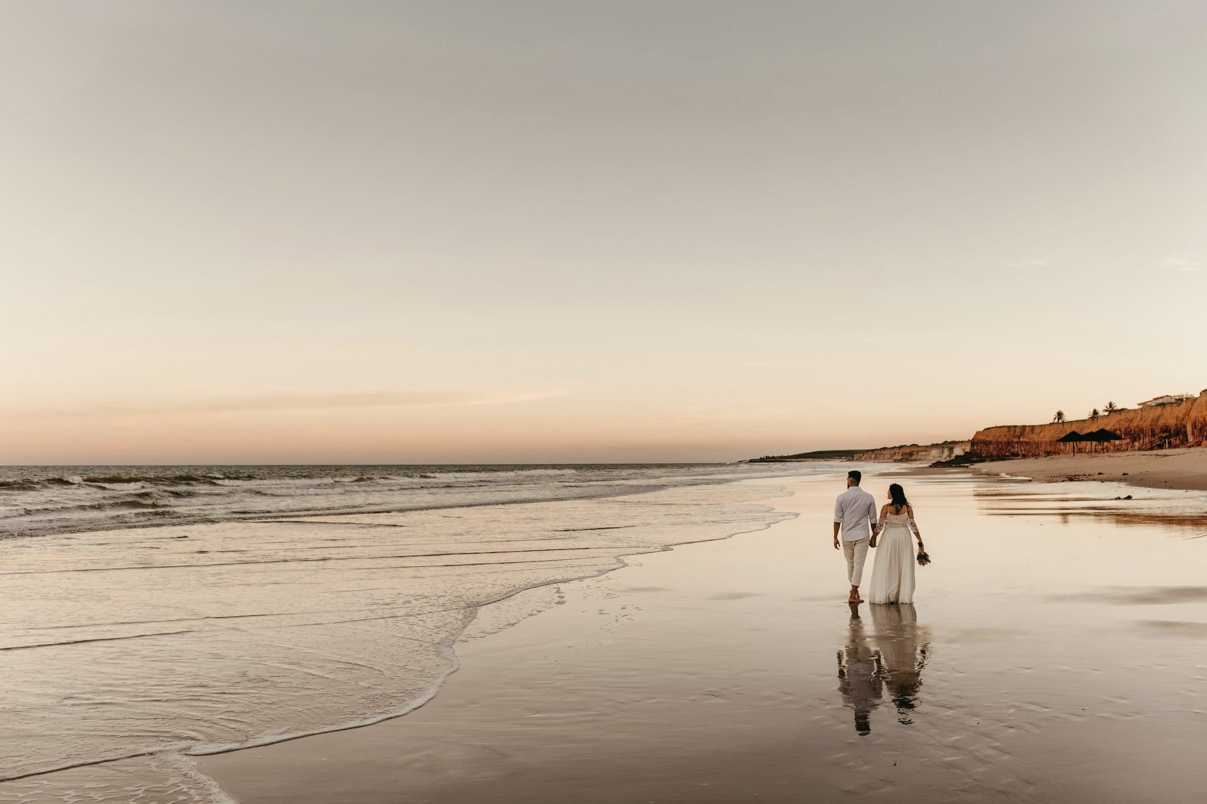 Pareja recién casada camina tomada de la mano por la orilla del mar al atardecer, con el cielo rosado reflejado en la arena mojada.