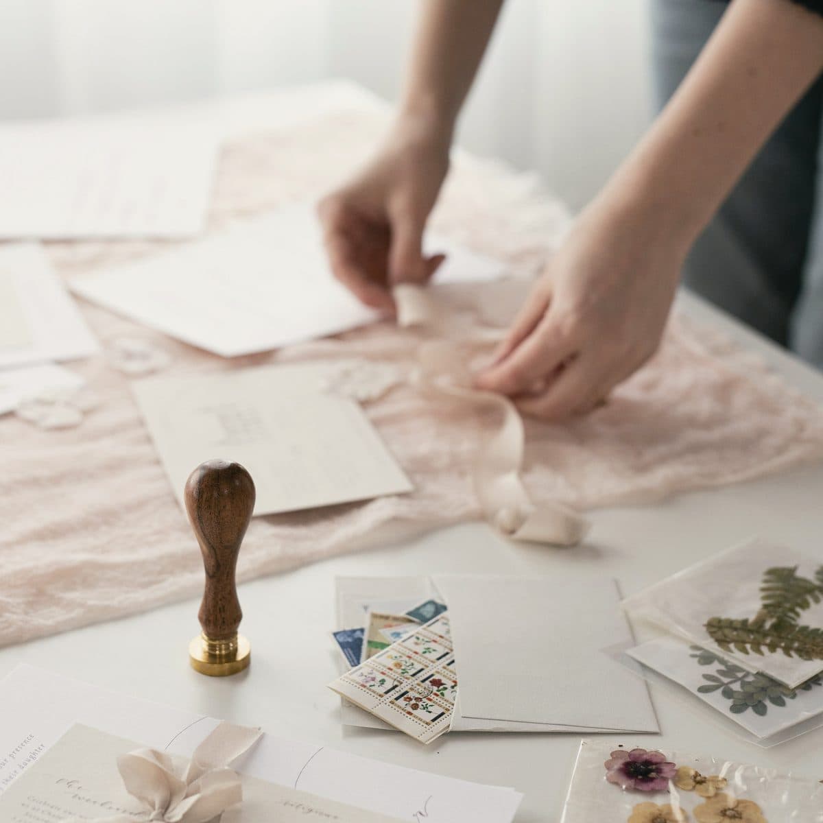 Manos organizando invitaciones, sello de lacre y muestras de tela sobre una mesa